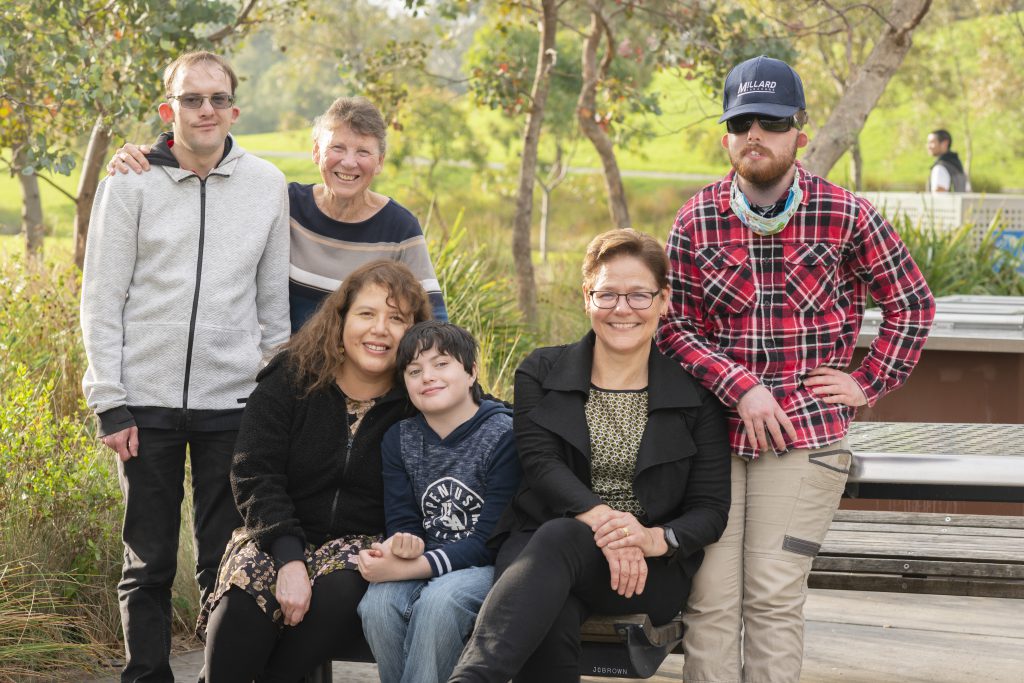 A group of consumer family members sit on a park bench and smile at the camera.