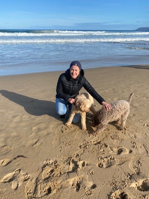 Adrienne is on a surf beach in winter wearing jeans, black puffer jacket and grey beanie, squatting down to pat Rosie and Freddie, the labradoodles.