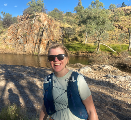 Alicia is smiling in her trail running gear as she takes a break from her trail run. She is surrounded by nature on the Larapinta Trail with a water hole, rocks and trees in the background.