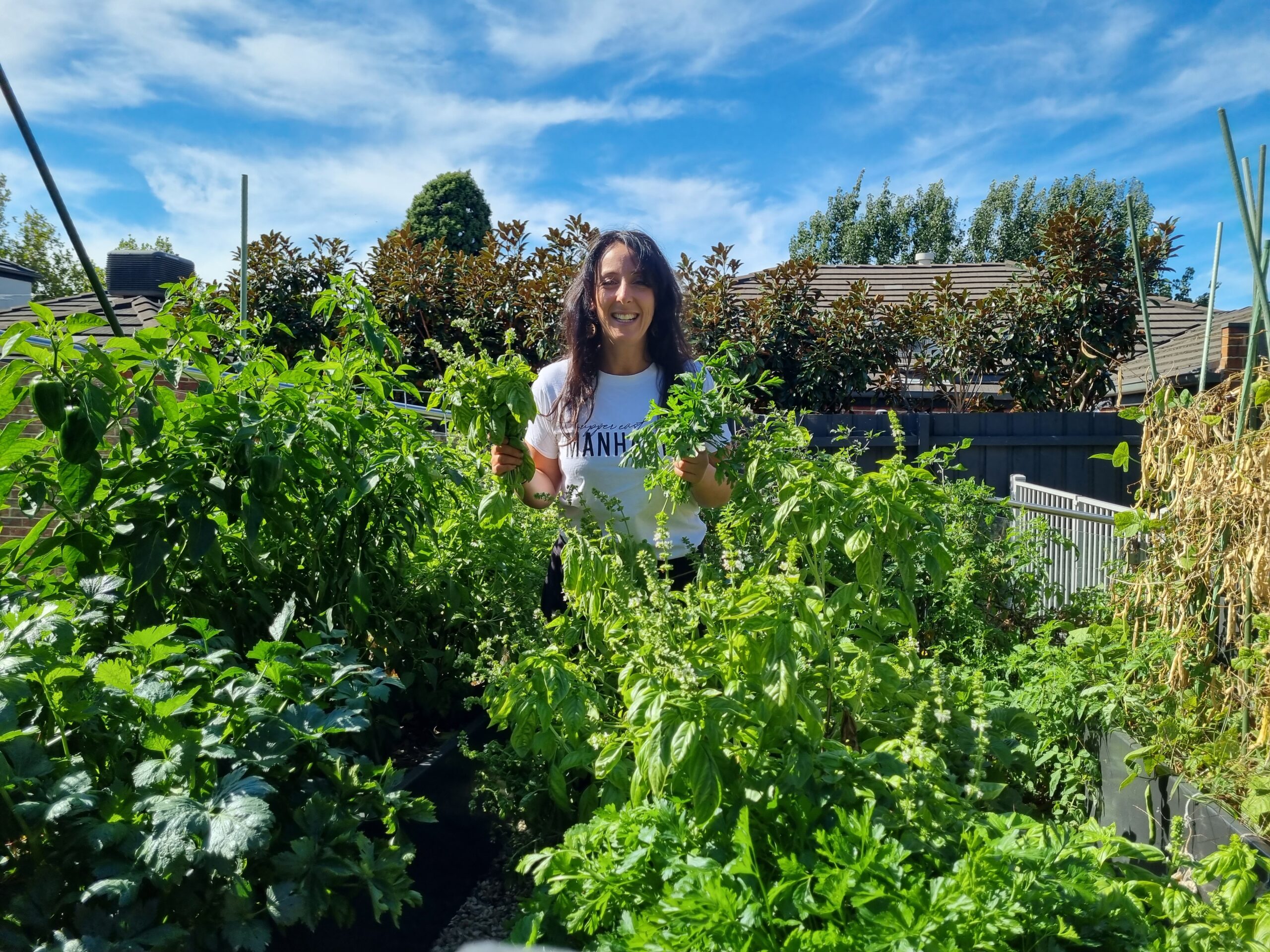 Catherine stands in a lush backyard veggie patch, in a white t-shirt with Manhattan written on the chest in black. She smiles happily holding up cuttings of basil in amongst shoulder height basil, capsicum, and tomato plants.