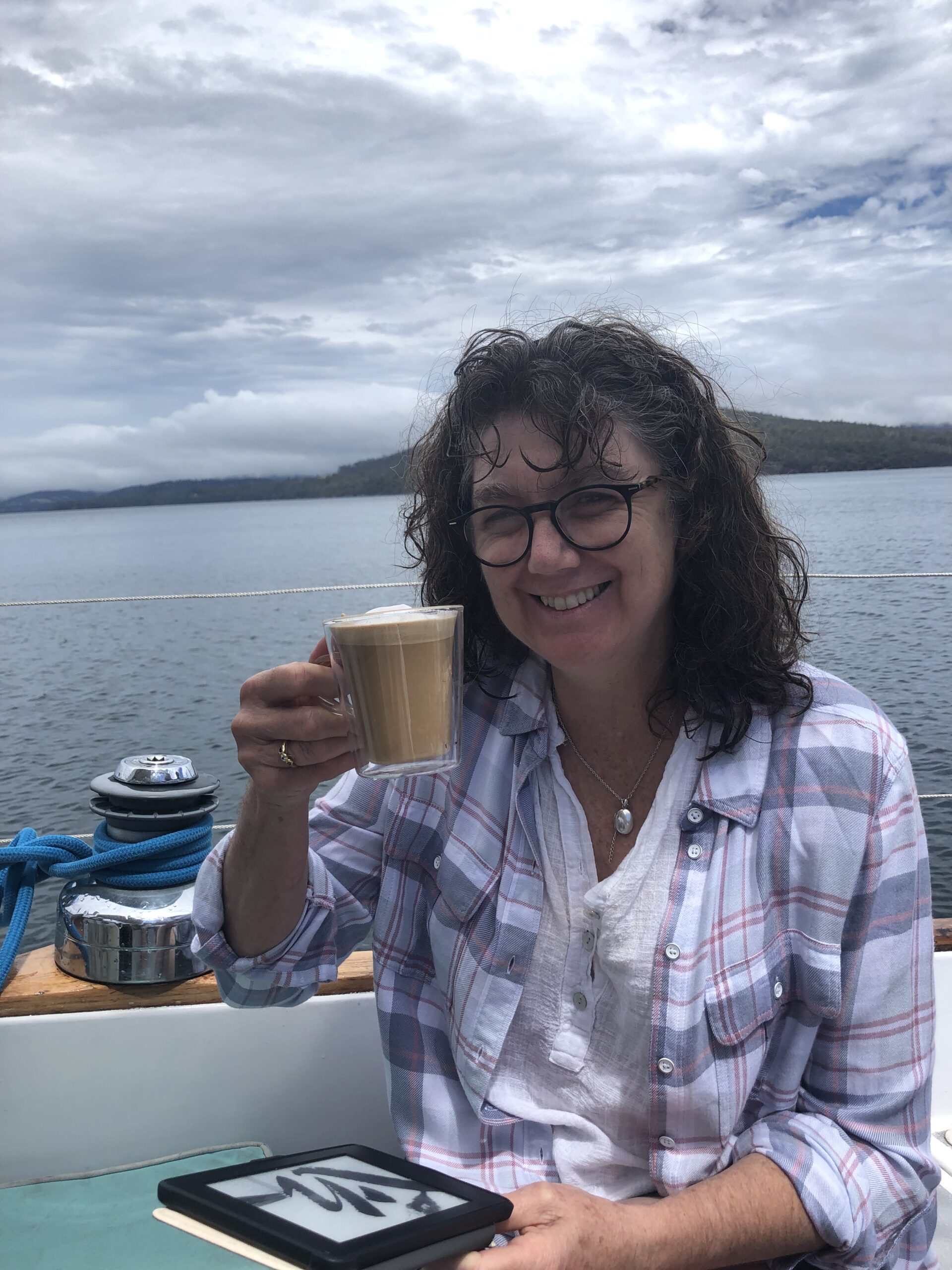 Christine is sitting on her boat drinking a coffee and reading her e-book. She has just had a swim, so her hair is wet and she is happy. The background is of smooth water and a hill in the background. The sky is cloudy.