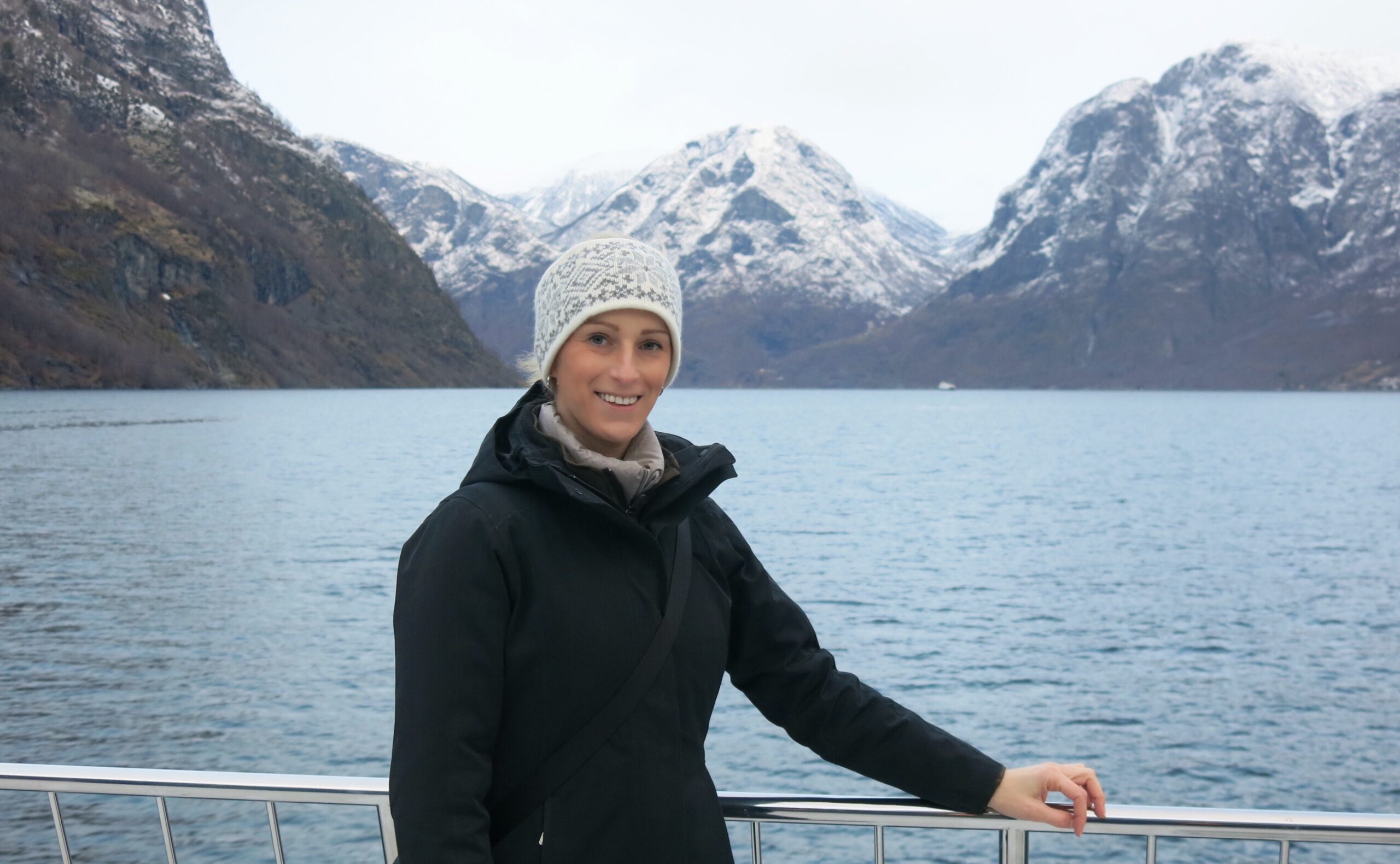 Georgie is standing at the bow of a boat, rugged up in a warm black jacket. She is on a cruise in Norway. The beautiful Aurlandsfjord and snow-covered mountains are in the background..