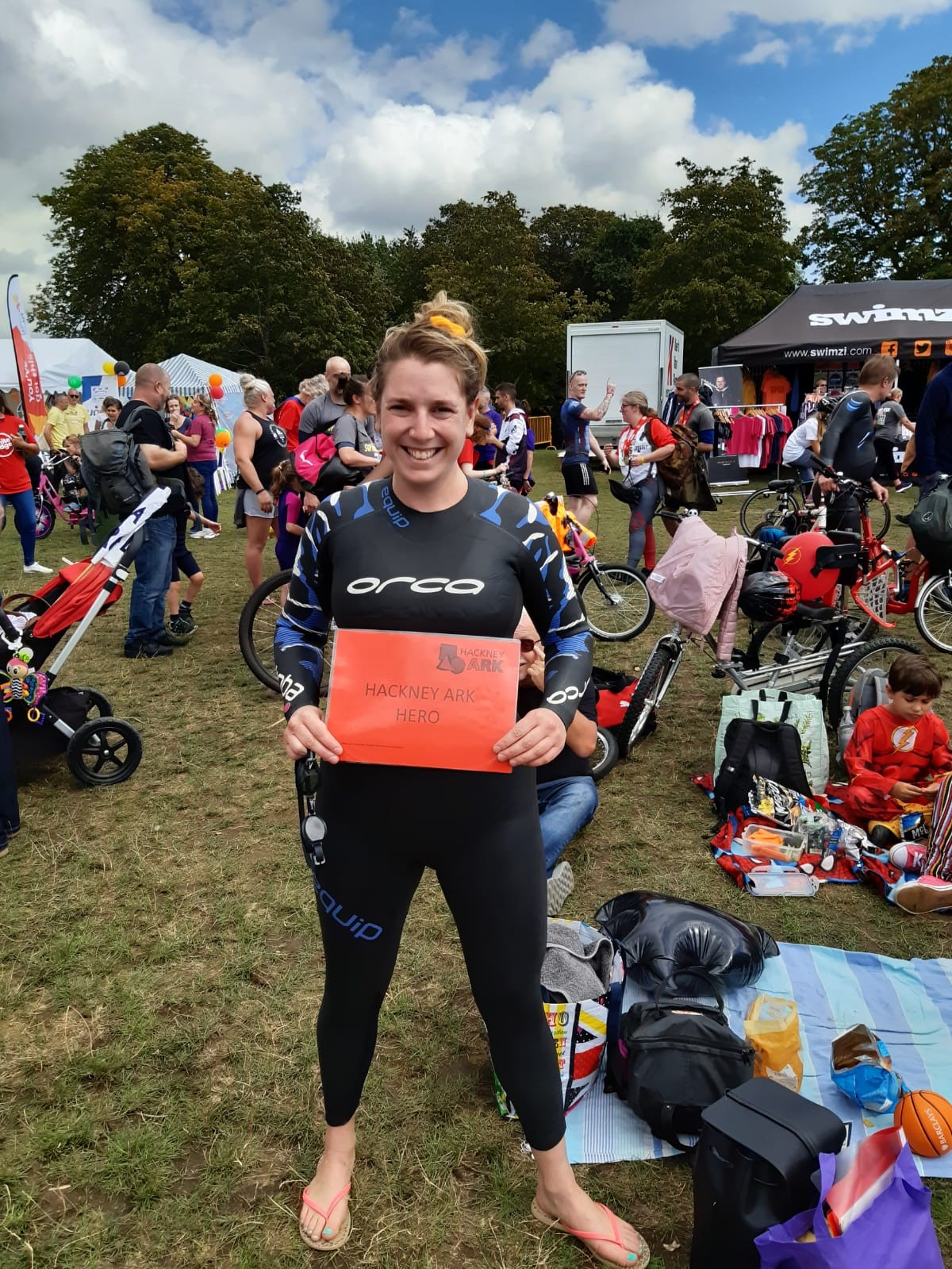 Jacky stands smiling in a wetsuit with a sign, ready to compete in the swim section of the Superhero Series triathlon. She is in a field surrounded by other families who have come to compete with many adapted bikes and prams in view