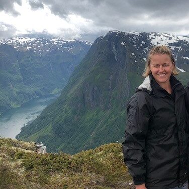 Kerry is standing at the top of a mountain overlooking a fjord in Norway. She is wearing a black raincoat and has wet hair. She is smiling because she hiked up in the rain, and made it to the top and the rain stopped