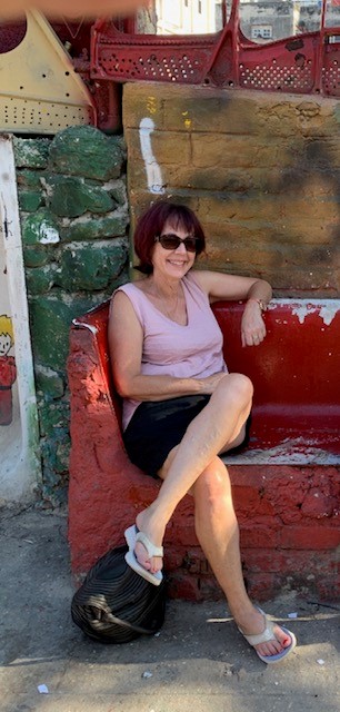 Vicki sits cross legged wearing a black skirt, pink singlet, thongs and sunglasses. She is smiling and relaxed sitting on a red brick bench.