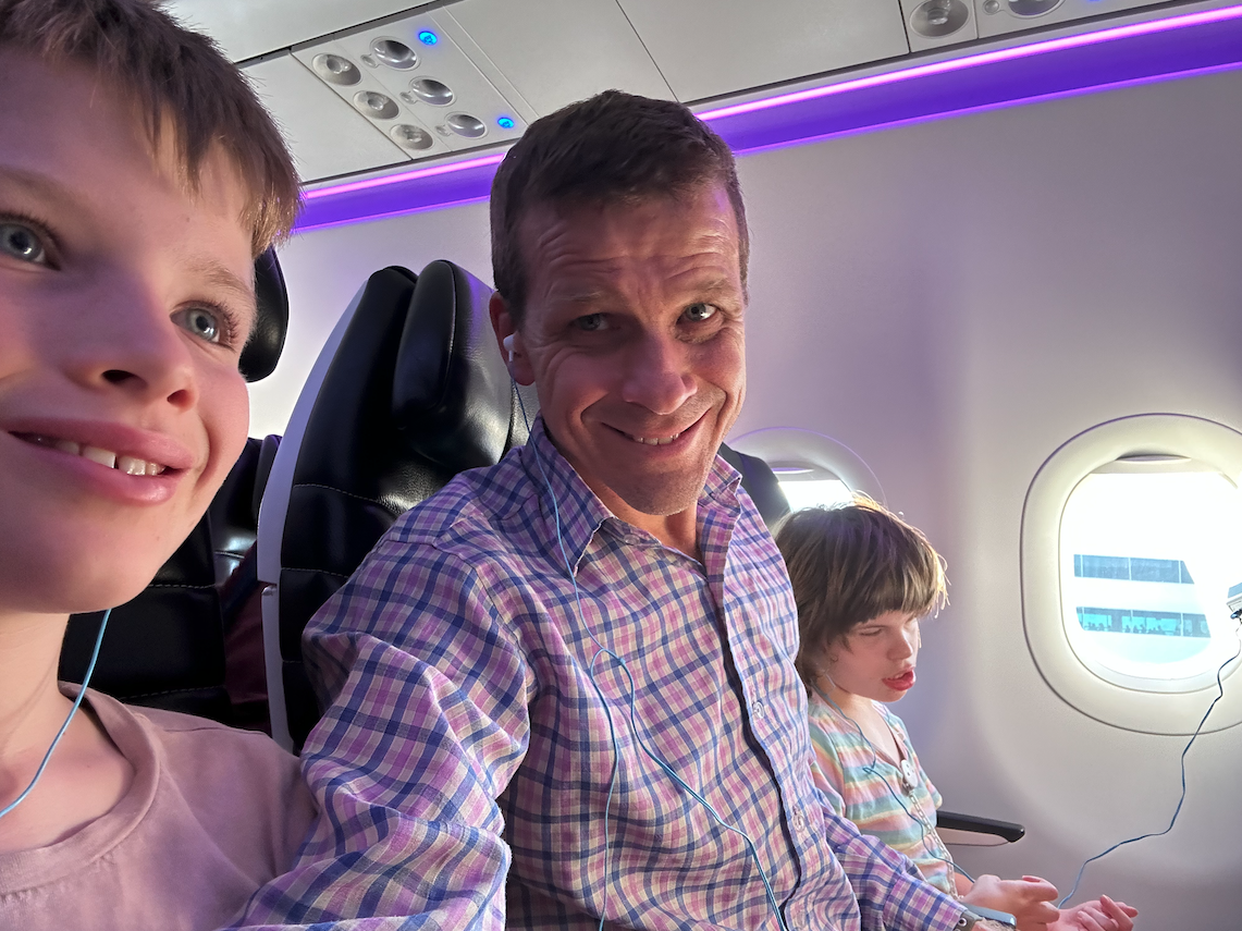Nathan is pictured wearing a check shirt, smiling at the camera and seated on a plane, between his daughter on his left and son on his right.