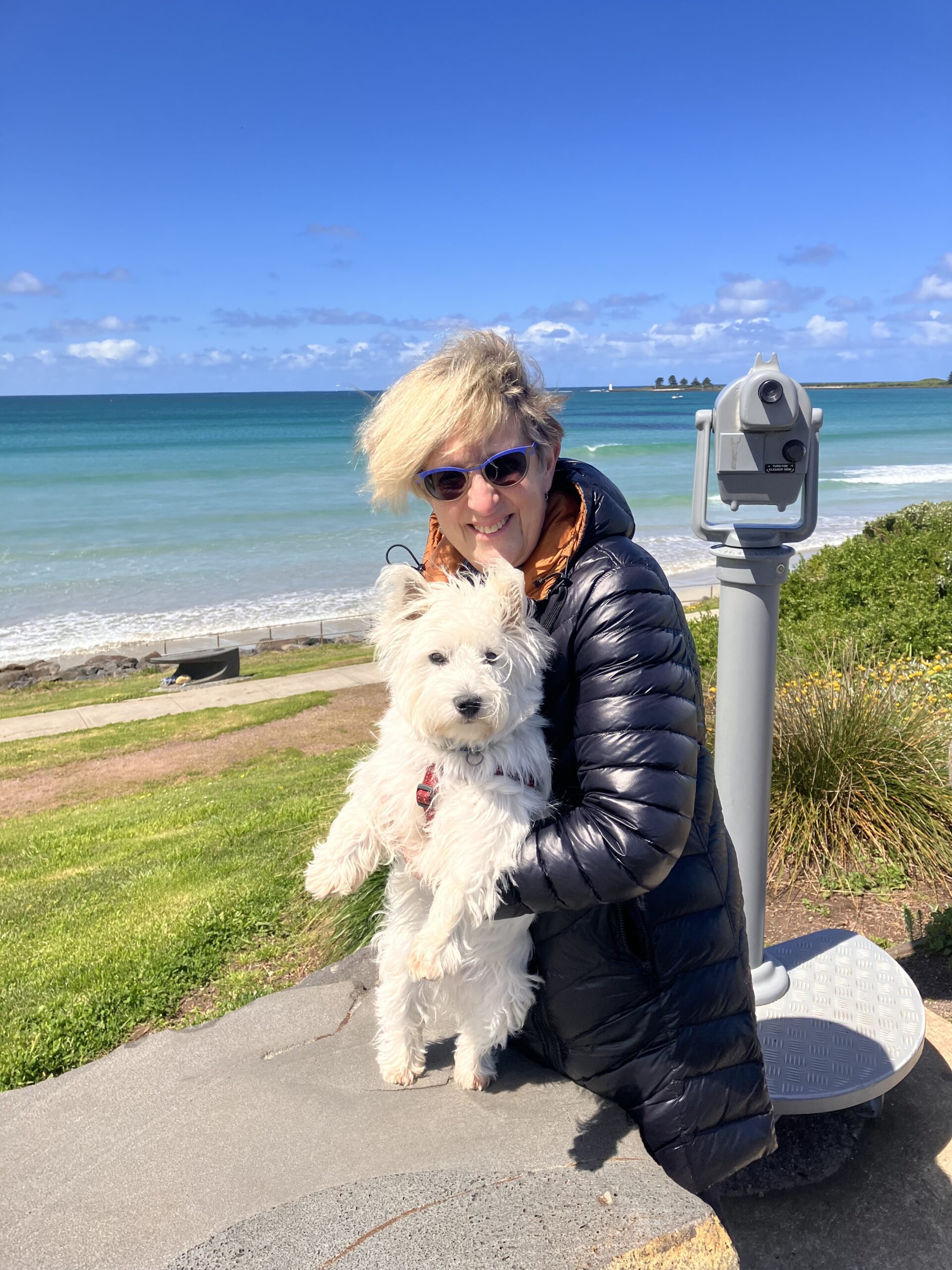 Sue is pictured at East Beach, Port Fairy, next to a lookout telescope, holding her 6 month-old white Westie puppy, Piper. She wears a black and orange overcoat and sunglasses.