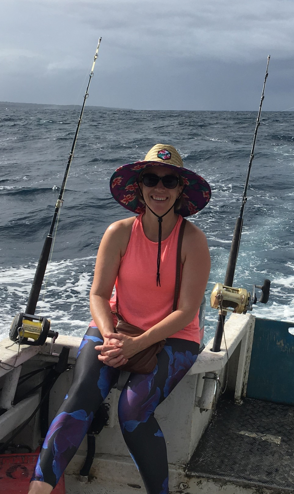 Nicole is smiling while sitting on a moving fishing boat between two large fishing rods. The beautiful Fiji ocean is in the background.