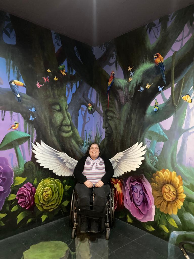 A picture of Freya smiling at the camera while sitting in a wheelchair. She is lined up with a colourful mural on the wall that makes it look like she has wings and is sitting in a forest.