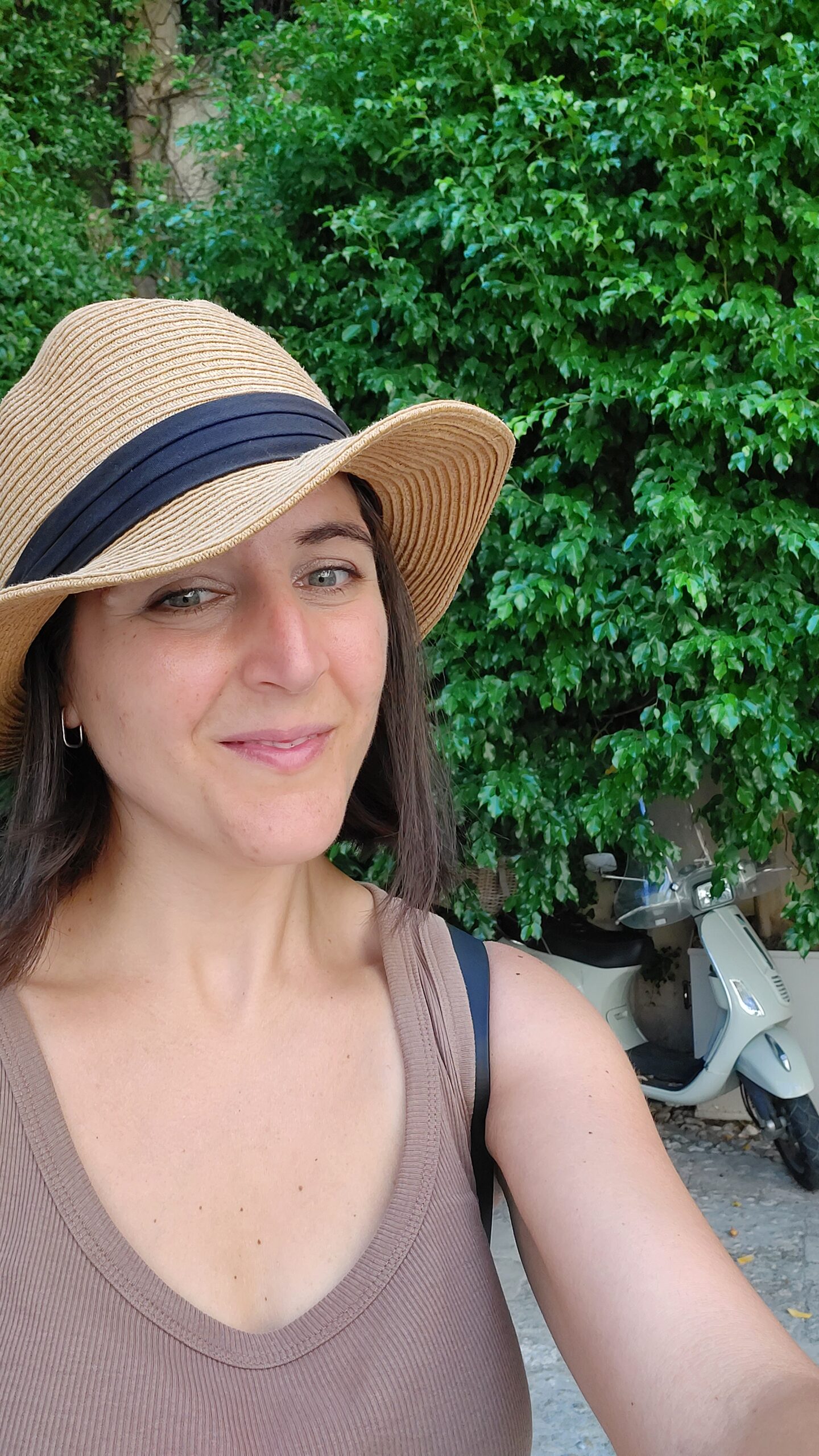 Francesca is smiling, wearing a hat, and standing in a leafy courtyard.