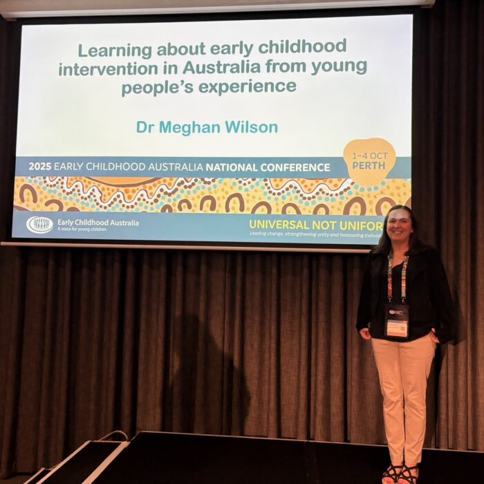 A picture of Meg Wilson standing in front of the opening slide of her presentation at the 2025 ECA national conference. The slide reads "Learning about early childhood intervention in Australia from young people's experience, Dr Meghan Wilson".